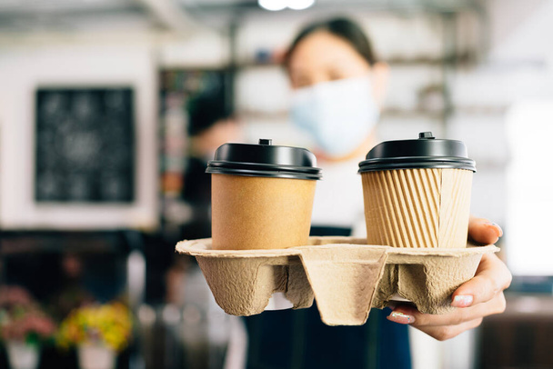 Woman Barista wearing a face mask serving coffee in takeaway paper disposable cups in the coffee shop. Small green business during coronavirus pandemic. Owner and employee service for the client. - Photo, Image