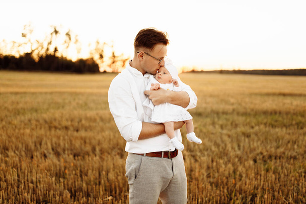 Handsome father with adorable little daughter at the field, caring dad hold newborn baby girl in arms, gently kiss cute child, parenting and fatherhood concept - Foto, afbeelding