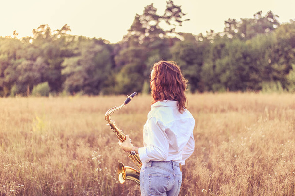 白いシャツを着た若い赤髪の女性が秋の野原に立って夕日を見ている。 - 写真・画像