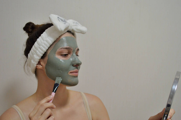 Ένα κορίτσι με επίδεσμο στο κεφάλι μουτζουρώνει μια πήλινη μάσκα στο πρόσωπό της ενώ κοιτιέται στον καθρέφτη. - Φωτογραφία, εικόνα
