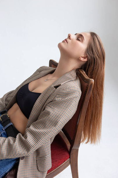 young caucasian woman in black top, jeans, suit jacket, lying on chair at studio - Photo, Image