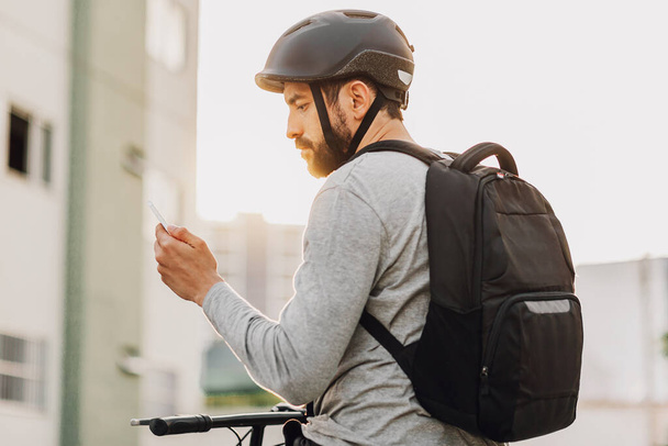 Νεαρός ενήλικας στέκεται στο ποδήλατο χρησιμοποιώντας smartphone στην πόλη - Φωτογραφία, εικόνα