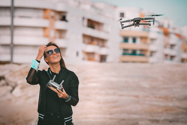 Die süße moderne lächelnde Pilotin in dunkler stylischer Kleidung steuert die Drohne per Fernbedienung. Hobby im Freien. - Foto, Bild