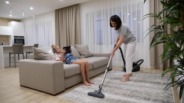 Moeder schoonmaken vloer in de woonkamer met een stofzuiger terwijl dochter luisteren muziek en het gebruik van tablet tijdens het zitten op de bank - Video