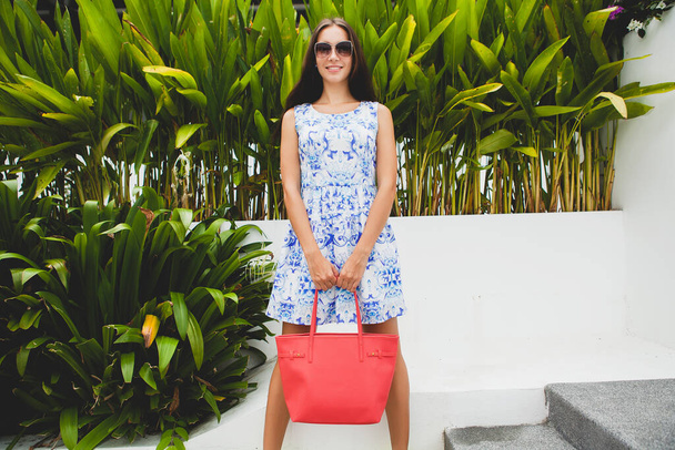 young stylish beautiful woman in blue printed dress, red bag, sunglasses, fashionable outfit, trendy apparel, smiling, sitting, summer, accessories - Фото, зображення
