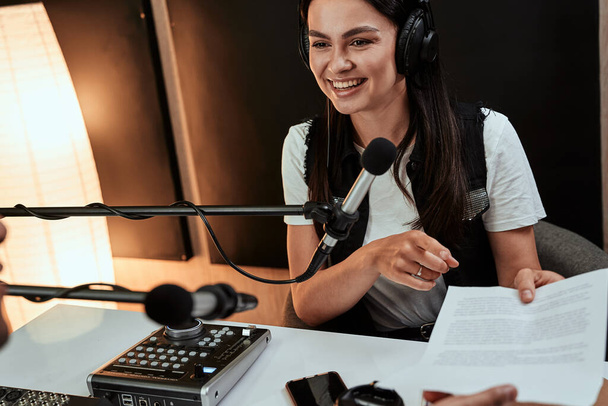 Portrait d'une animatrice radiophonique heureuse souriant et donnant un script papier à son collègue masculin tout en animant une émission en direct en studio - Photo, image
