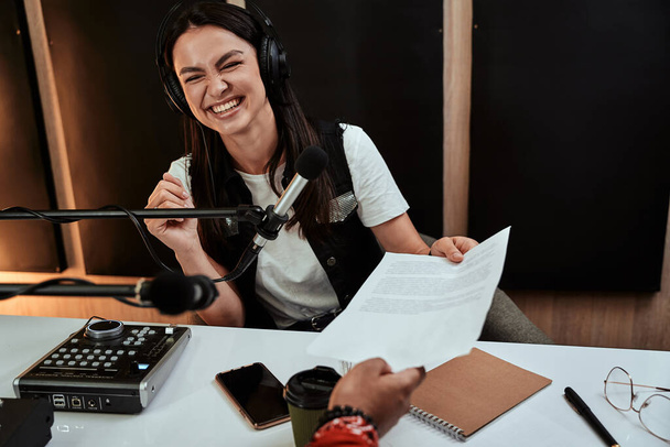 Portait de la feliz presentadora de radio femenina sonriendo, recibiendo un papel de guion de su colega masculino mientras modera un espectáculo en vivo en el estudio - Foto, imagen