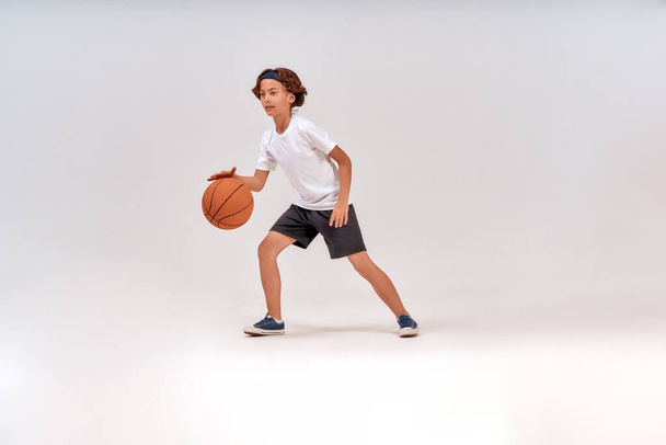 Çocuklar için en iyi spor. Gri arka planda tek başına duran, basketbol oynayan bir çocuğun tam boy çekimi. - Fotoğraf, Görsel