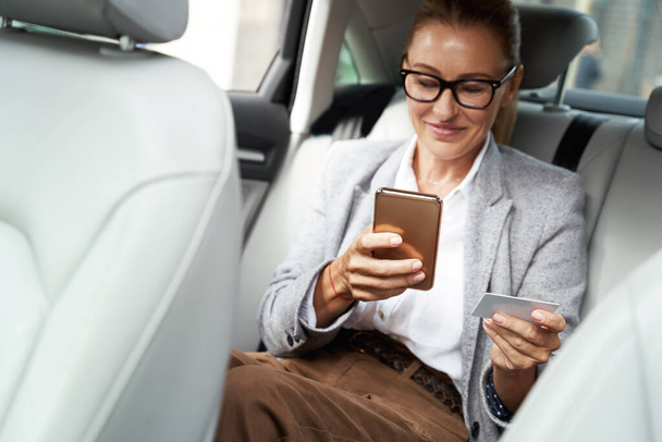 Щаслива успішна бізнес-леді в окулярах, використовуючи свій смартфон і кредитну картку, щоб купити щось, сидячи на задньому сидінні в машині
 - Фото, зображення