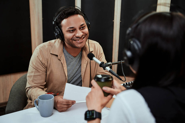 Portret van de jonge mannelijke radio-presentator live in de ether, in gesprek met een vrouwelijke gast, met een scriptpapier terwijl hij in de studio zit - Foto, afbeelding