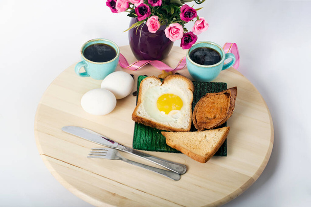 Desayuno en tabla de cortar de madera aislado sobre fondo blanco con camino de recorte. Huevo frito con tostadas en tazón de cerámica, dos tazas de espresso, huevos cocidos y hermosas flores moradas en jarrón - Foto, Imagen