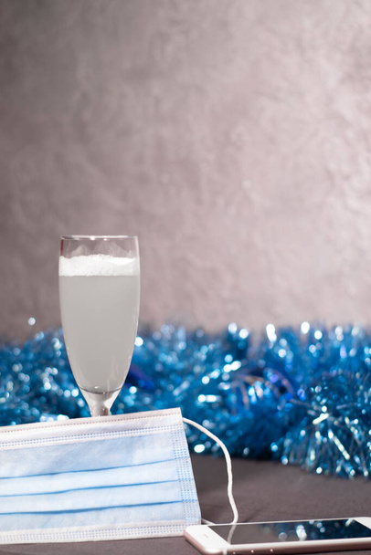 glass of champagne near a blue medical mask, phone and blue shiny tinsel. distance celebration. pandemic coronavirus - Photo, Image