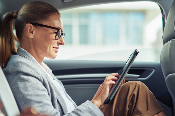 車の後部座席に座っている間、デジタルタブレットを使用して眼鏡をかけて成功した笑顔のビジネス女性の側面 - 写真・画像