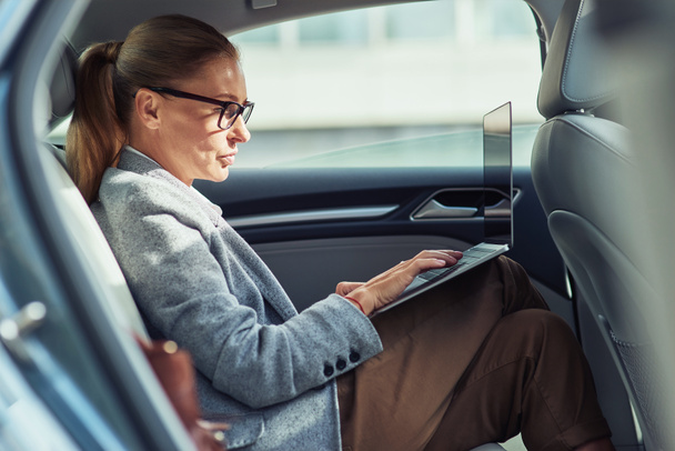 Sprawdzam e-mail. Widok z boku skupienia bizneswoman w średnim wieku za pomocą laptopa podczas siedzenia na tylnym siedzeniu w samochodzie - Zdjęcie, obraz