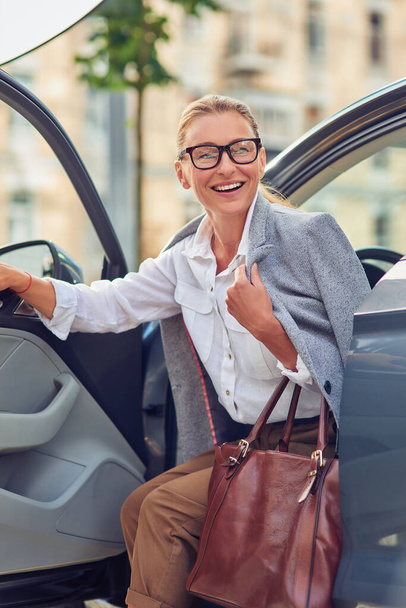 Успішна модна бізнес-леді середнього віку з сумочкою, що виходить з її сучасної машини і посміхається, приїхала на роботу
 - Фото, зображення