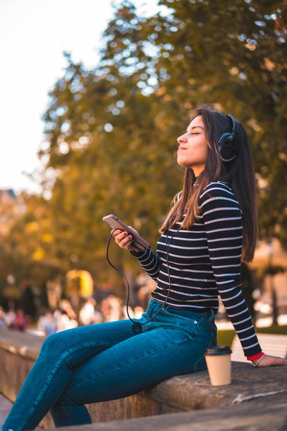 Lifestyle, Caucasian brunette enjoying listening to music in the city. Relaxing with a take away coffee jungo to a beautiful church in the park. Black sweater with white stripes - Photo, Image
