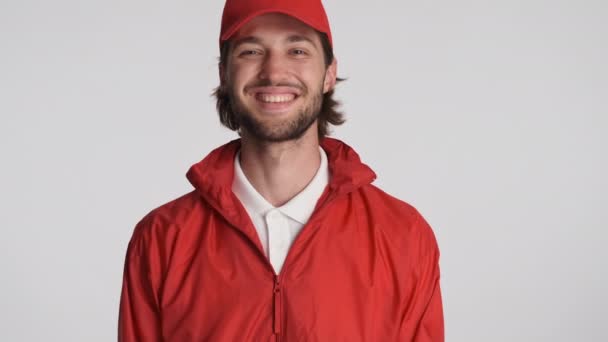 Portrait du livreur souriant en casquette rouge et veste regardant joyeusement à la caméra et agitant oui sur fond blanc - Séquence, vidéo