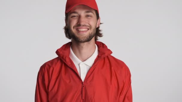 Atractivo repartidor en gorra roja y chaqueta felizmente mirando en cámara y el pulgar hacia arriba gesto sobre fondo gris. Expresión bien hecha - Imágenes, Vídeo
