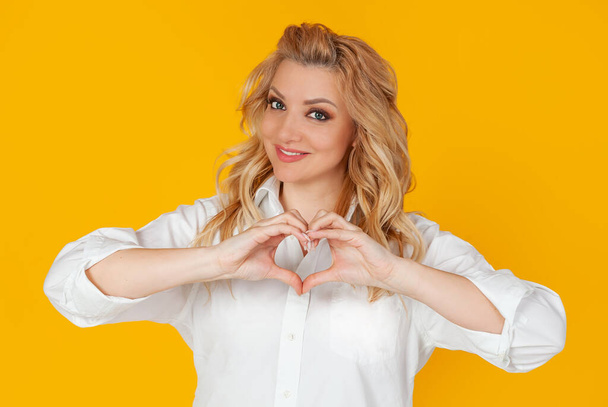 Blondine woman in white shirt, middle aged European girl, showing heart gesture, recognizing sympathy, look smiling friendly at the camera, girlfriend expresses true feelings, on yellow background. - Фото, зображення