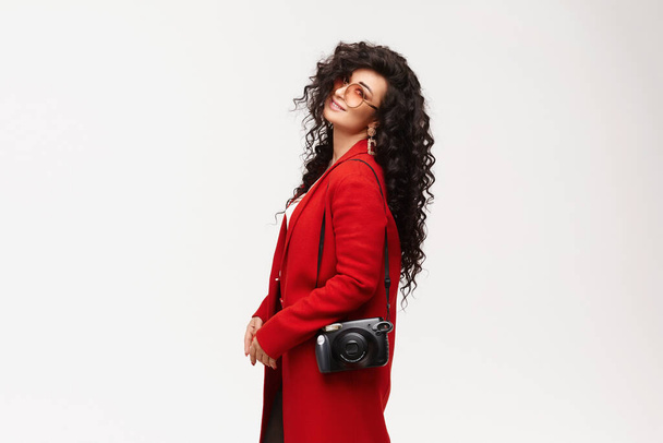 Молодая модель девушка с черными вьющимися волосами в красном пальто и круглые очки, позирующие боком, изолированные на белом. - Фото, изображение