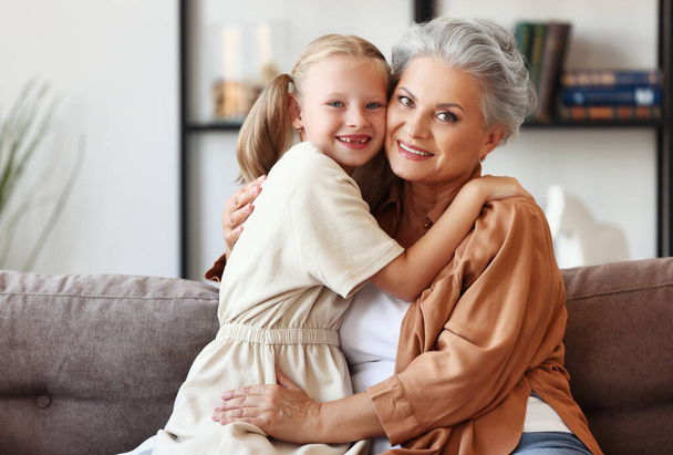 Radostná holčička objímající šťastnou babičku, zatímco sedí spolu na pohovce a dívá se na kameru na prarodiče da - Fotografie, Obrázek
