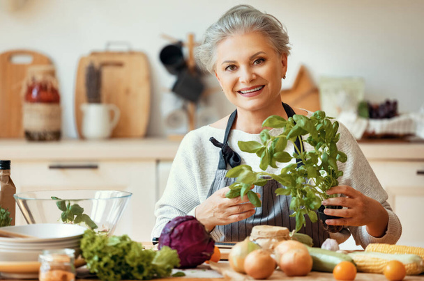 Vidám középkorú háziasszony kötényben, kezében egy csomó friss zöld bazsalikommal, és a kamerába néz, miközben egészséges ételt készít friss zöldségekkel az otthoni konyhában. - Fotó, kép