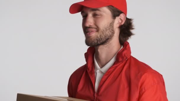 Hombre de entrega guapo en gorra roja felizmente dando caja de entrega en la cámara sobre fondo blanco - Imágenes, Vídeo