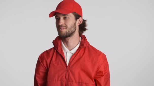 Attractive courier man in red cap happily catching delivery box on camera over white background. Service concept - Imágenes, Vídeo