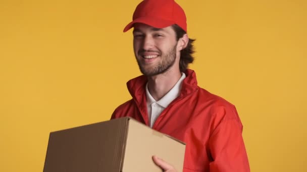 Fešák kurýr v červené čepici radostně dává doručovací box na kameru přes barevné pozadí. Šťastný výraz - Záběry, video