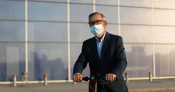 Senior Caucasian good-looking man in glasses and medical mask riding electric scooter at modern big glass building at aeroport. Grandfather having nice ride outdoor in urban city. Pandemic concept. - Foto, Imagem
