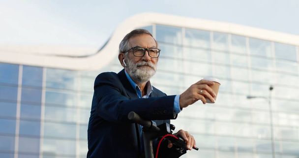 Senior gray-haired man in glasses and headphones standing at bike on street and drinking coffee to-go. Old grandfather in airpods listening to music and sipping hot drink outdoor. - Photo, Image