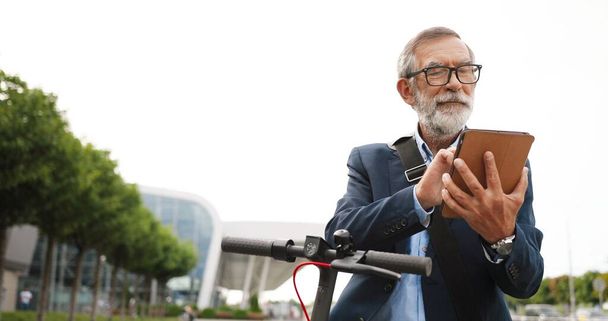 Senior gray-haired man in glasses and headphones standing at bike on street and tapping or scrolling on gadget computer. Old grandfather in eyeglasses using tablet device and browsing online outdoor. - Photo, Image