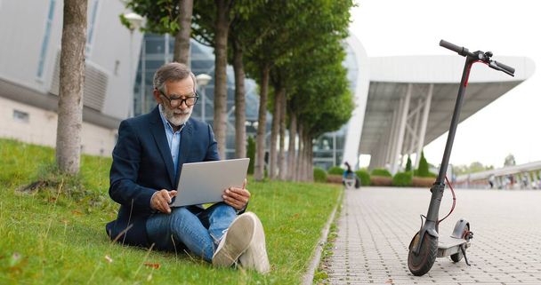 Old Caucasian man in glasses sitting outdoor on green grass at electric scooter and working on laptop computer. Male typing on keyboard at street. Grandfather browsing online. Businessman pensioner. - Foto, immagini