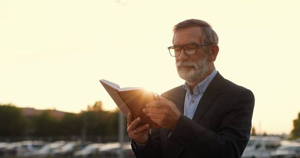 Old Caucasian gray-haired man in glasses walking and reading book outdoor in sunset at cars parking on background. Senior male professor strolling and read textbook. Studying. - Photo, image
