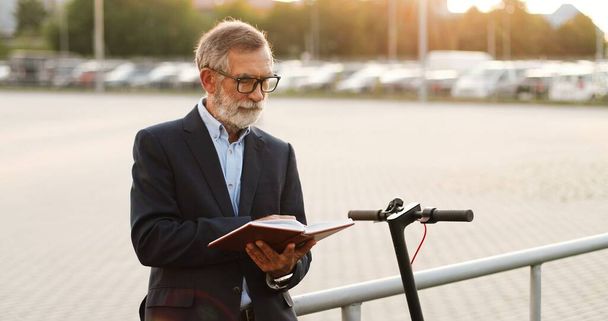 Old Caucasian gray-haired man in glasses walking and reading book outdoor in sunset at cars parking on background. Senior male professor strolling and read textbook. Studying. - Foto, immagini