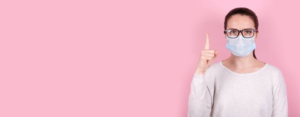 Portrait of a brunette girl with eyeglasses in a medical mask showing finger up, idea concept. Isolated on pink background. - Fotografie, Obrázek