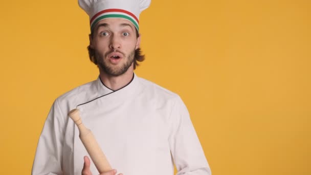 Attraente chef professionista italiano in uniforme mostrando emotivamente bellissimo gesto e mattarello sulla macchina fotografica su sfondo colorato - Filmati, video