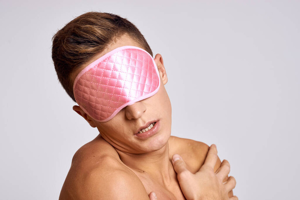 Un hombre con una máscara rosa para dormir sobre un fondo claro por la noche gestos con las manos - Foto, Imagen