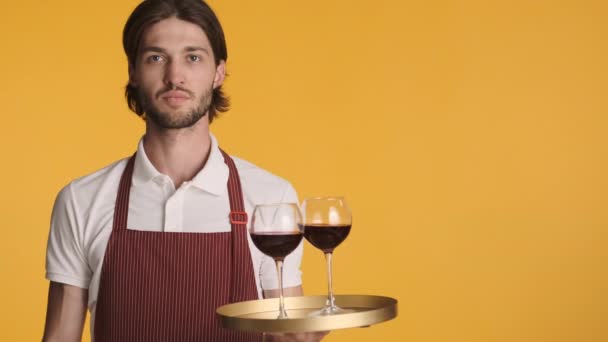 Joven camarero alegre atractivo sosteniendo bandeja con vino tinto sonriendo alegremente en la cámara sobre fondo colorido - Metraje, vídeo