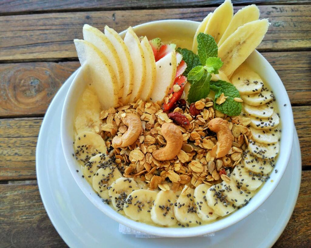 Healthy fruit smoothie bowl topped with fresh bananas, Asian pear, cashews, chia seeds, granola and a sprig of mint on a wooden table background - Photo, Image