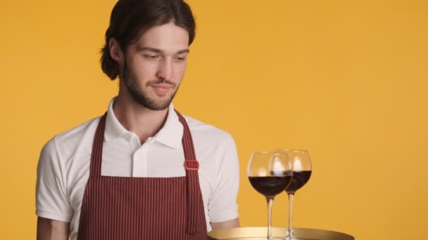 Joven camarero barbudo atractivo sosteniendo bandeja con vino tinto sonriendo alegremente en la cámara sobre fondo colorido - Metraje, vídeo