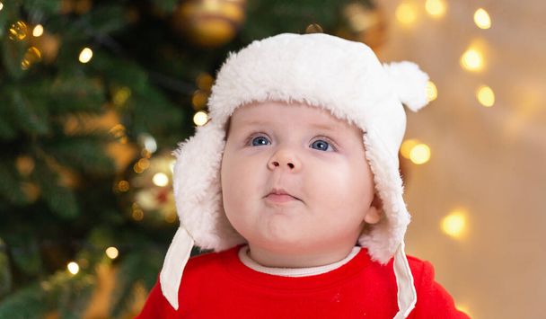 Karácsonyi gyerek figyelmes, álmomban meglepett érzelmek. Egy csinos kislány piros ruhában és fehér kalapban kifejezi az érzelmeit. Karácsonyi koncepció kisgyerek fa és koszorú a háttérben elmosódott - Fotó, kép