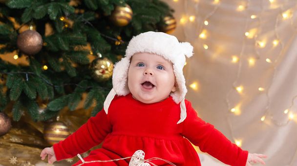 Karácsonyi gyerek nyugdíjasan, álomszerűen felnéz. Egy csinos kislány piros ruhában és fehér kalapban kifejezi az érzelmeit. Karácsonyi koncepció kisgyerek, fa és koszorú a háttérben elmosódott - Fotó, kép