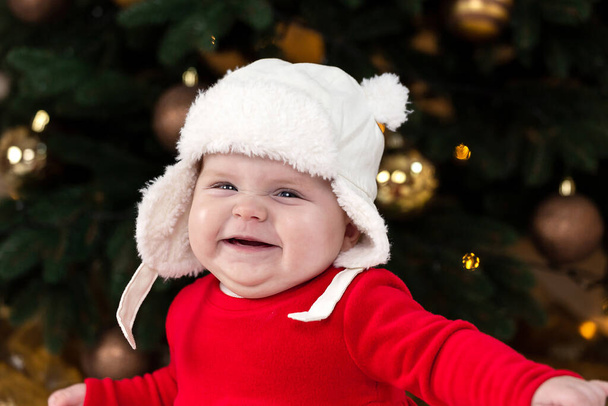 Jouluvauva hymyilee ja irvistää. Söpö pieni tyttö punaisessa mekossa ja valkoisessa hatussa ilmaisee tunteita. Joulukonsepti pieni lapsi, puu ja seppele taustalla hämärä - Valokuva, kuva