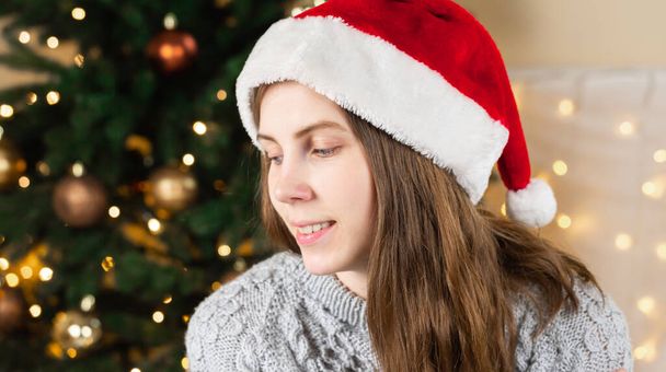 Portrait of a young woman in a gray sweater and santa claus hat sits near a Christmas tree against the background of a glowing garland.  - Photo, image