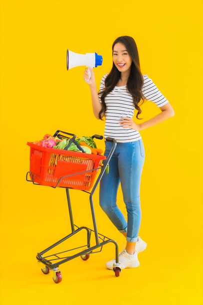 Porträt schöne junge asiatische Frau mit Einkaufswagen und Megafon auf gelbem Hintergrund - Foto, Bild