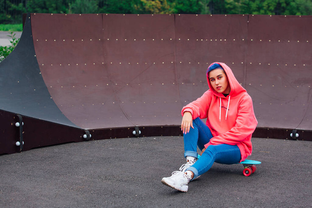 Stylish young woman sitting on her plastic skateboard in skatepark. Youth concept. - Фото, изображение