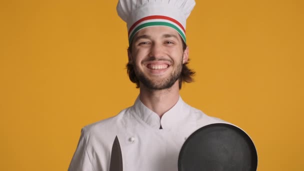 Chef sonriente profesional en uniforme que sostiene el cuchillo y la sartén con entusiasmo mirando en cámara sobre fondo colorido - Imágenes, Vídeo