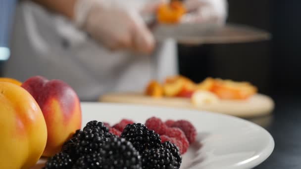 Cocinar ensalada fresca y fruta flamenca. Imagen borrosa de manos de chef rebanando jugoso albaricoque disparado a través de bayas en el plato. En cámara lenta. Concepto de alimentación saludable. Comida de restaurante. Full hd - Metraje, vídeo