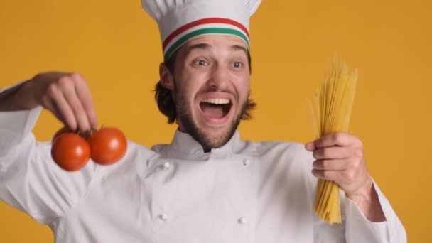 Atractivo chef italiano alegre en uniforme sosteniendo tomates y macarrones mirando alegremente en cámara sobre fondo colorido - Imágenes, Vídeo